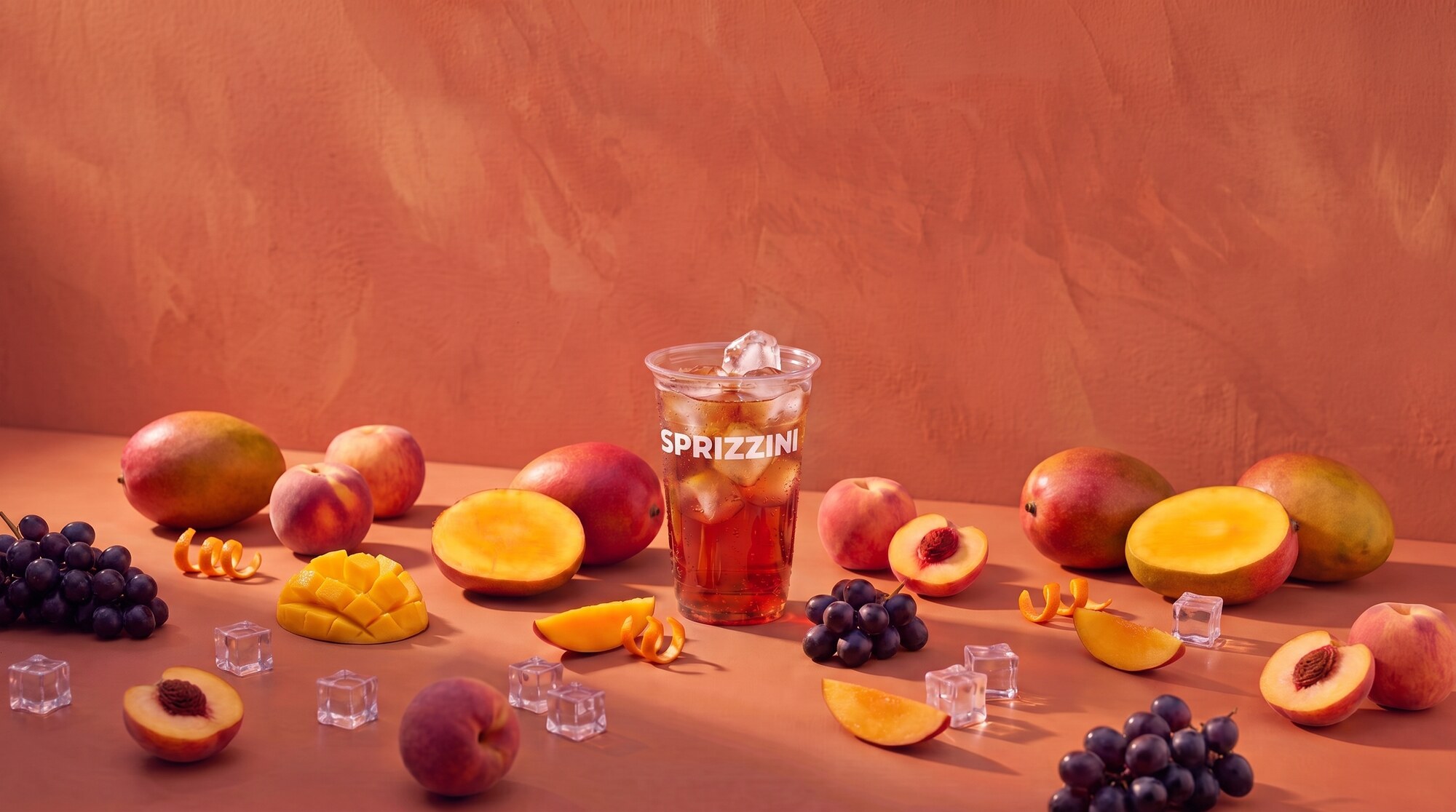 A Mango Lambrusco in a branded Sprizzini cup on a terracotta studio set, mango, peach and grapes around it